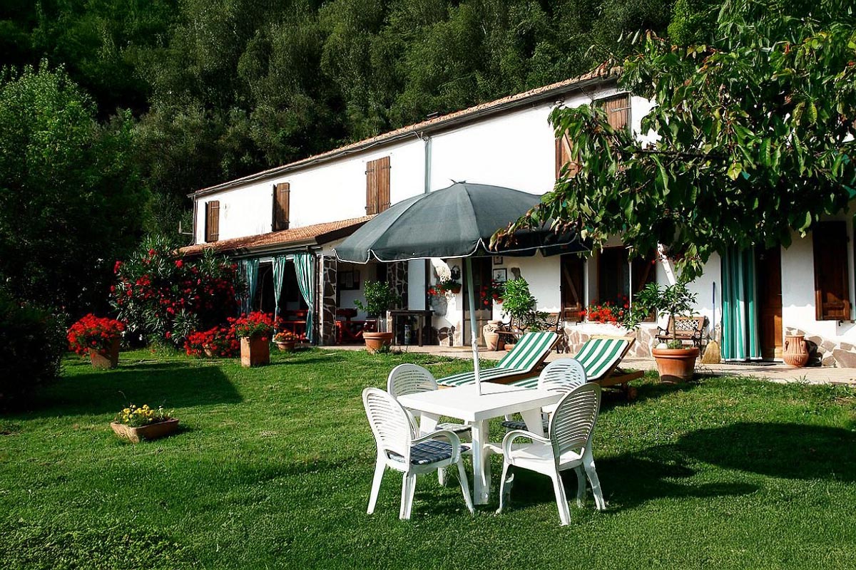 Weiße Fassade des Agriturismo La Bianca Fattoria mit grünen Fensterläden, Liegestühlen, Sonnenschirm und blühendem Garten vor der Kulisse bewaldeter Hügel.
