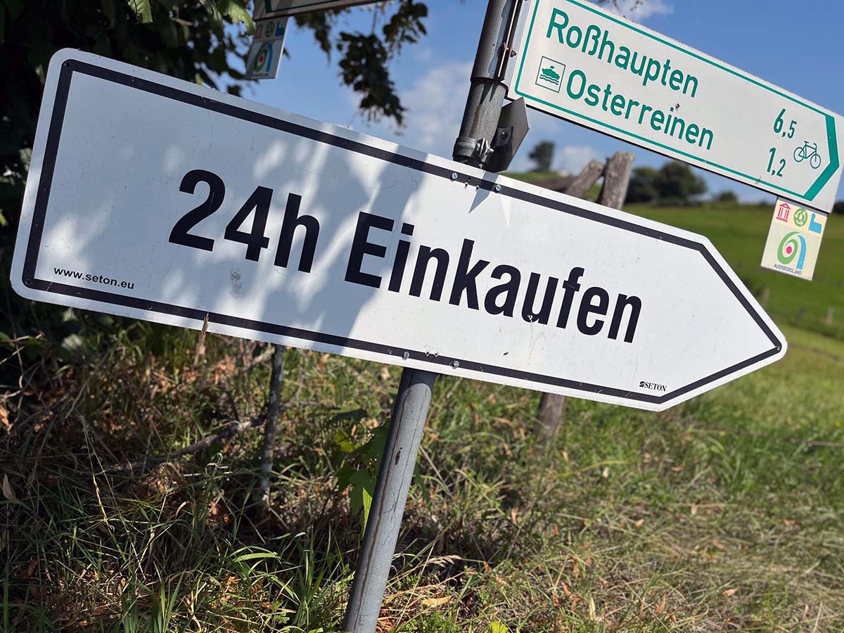 Wegweiser mit der Aufschrift „24h Einkaufen“ an einer Landstraße, Symbol für Hofläden und Milchtankstellen in Deutschland mit regionalen Produkten direkt vom Bauernhof.