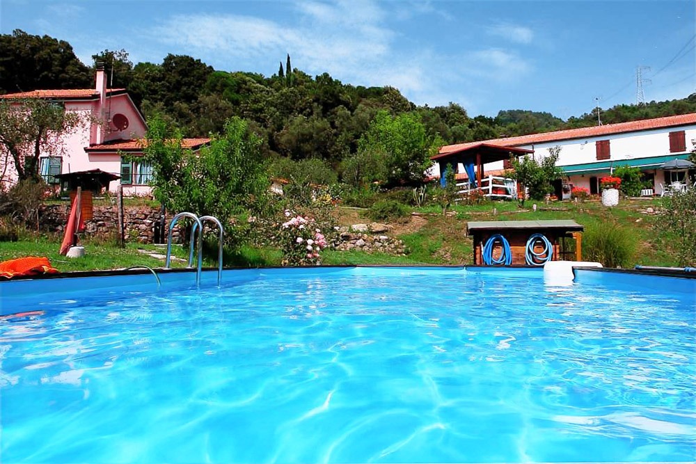 Blauer Außenpool des Agriturismo La Bianca Fattoria, umgeben von Garten, Blumen und den weißen Gebäuden der Fattoria mit roten Fensterläden.