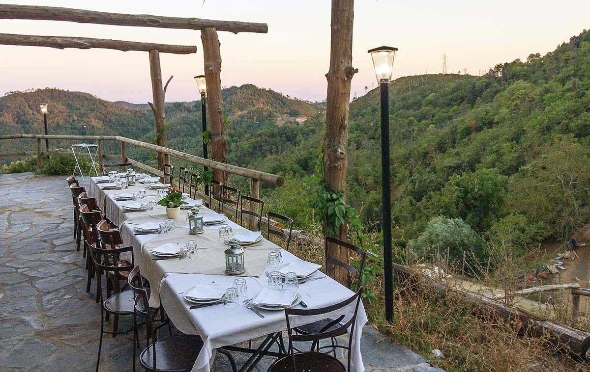 Lange Tafel auf der Restaurantterrasse der Agriosteria des Agriturismo degli Olivi, liebevoll gedeckt für Gäste, umgeben von Hügeln und Wäldern, im warmen Licht des Sonnenuntergangs.