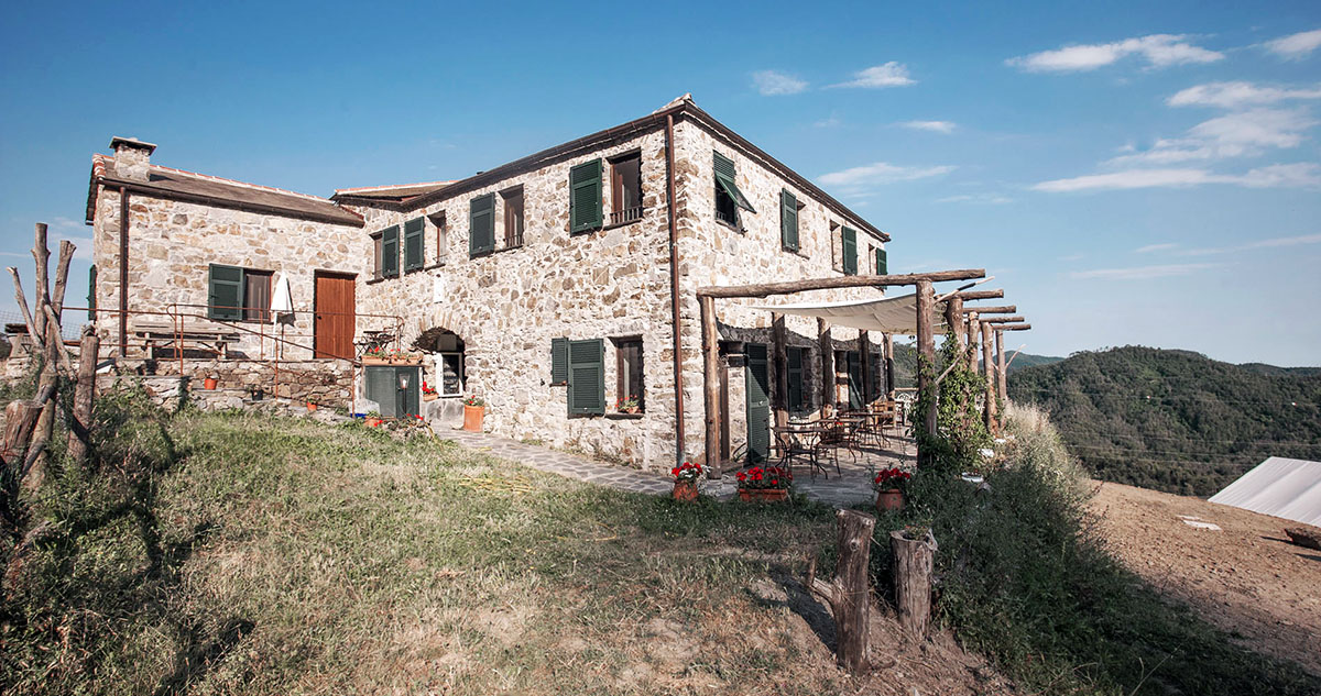Steinhaus des Agriturismo degli Olivi in Ligurien, umgeben von Hügeln und Natur, mit überdachter Terrasse, Holzmöbeln und blühenden Blumen vor dem Eingang. Im Hintergrund weiter Blick auf das ligurische Hinterland unter blauem Himmel.