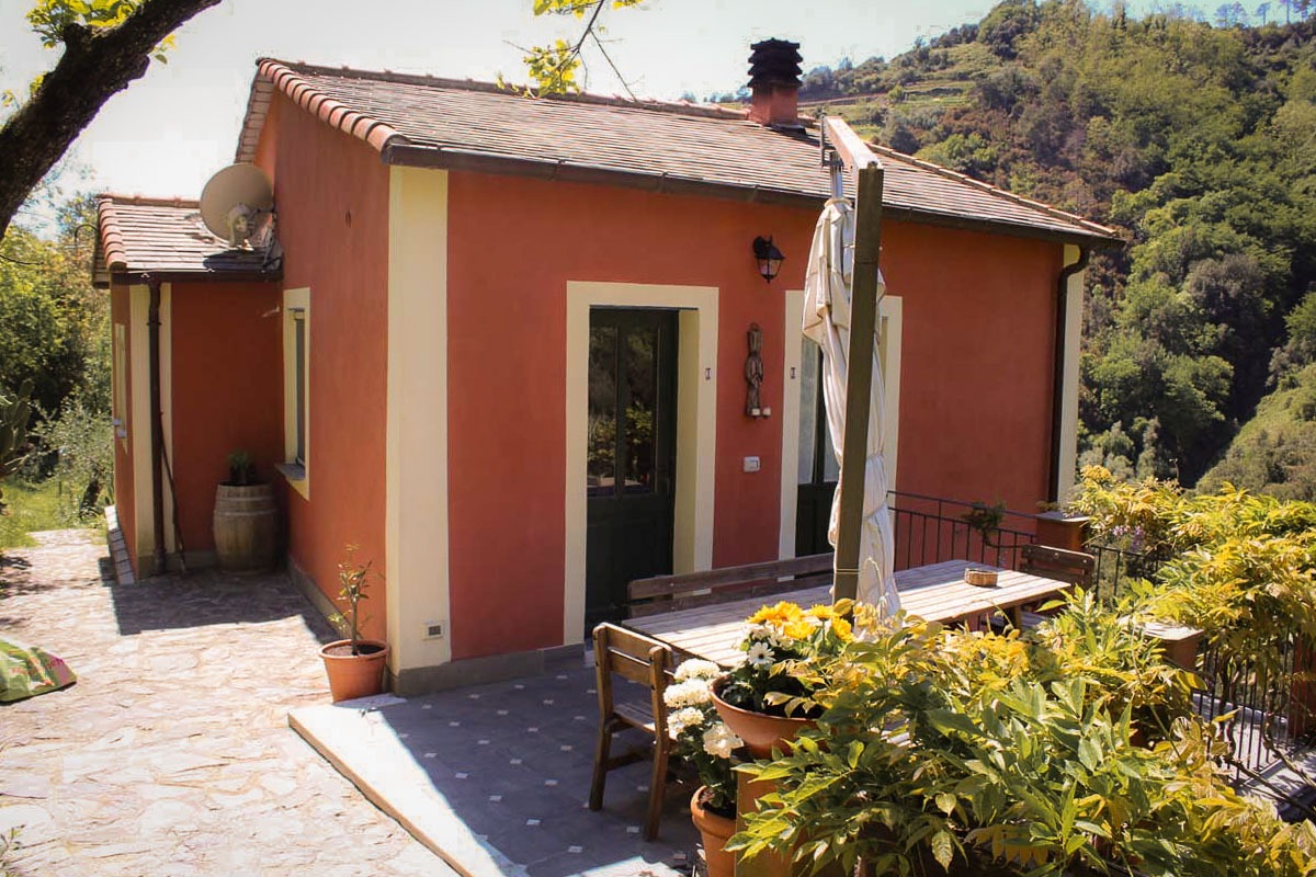 Terrakottafarbenes Gebäude des Agriturismo Il Brucaolivo mit grünen Türen, blühenden Pflanzen und Terrasse, umgeben von den Hügeln der Cinque Terre.