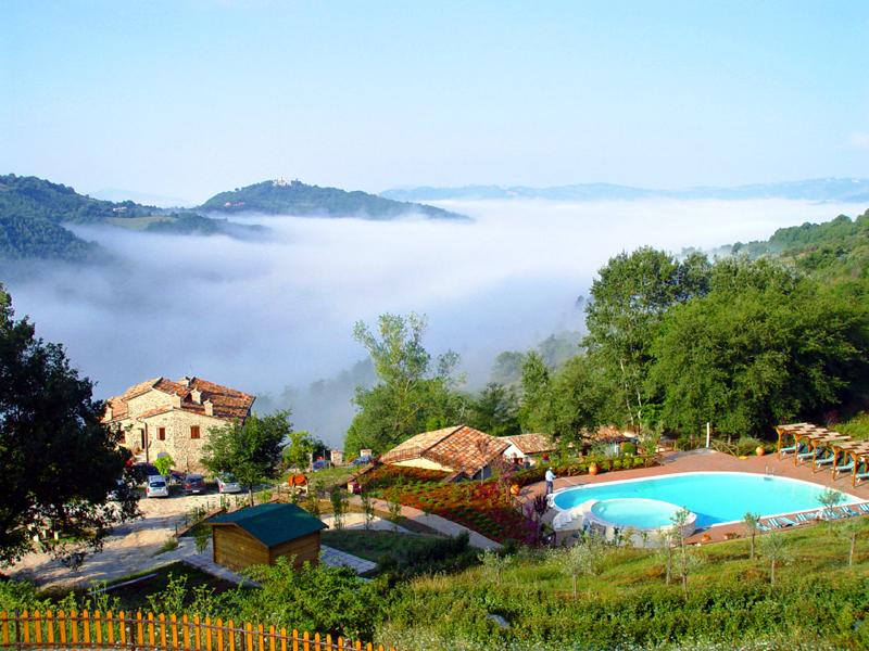 Blick von oben auf das Agriturismo Val di Boccio in Umbrien: Im Vordergrund das Natursteinhaus und der Pool, dahinter grüne Hügel und ein Nebelmeer, das sich sanft durch das Tal zieht – eine friedliche, malerische Landschaft inmitten der Natur.