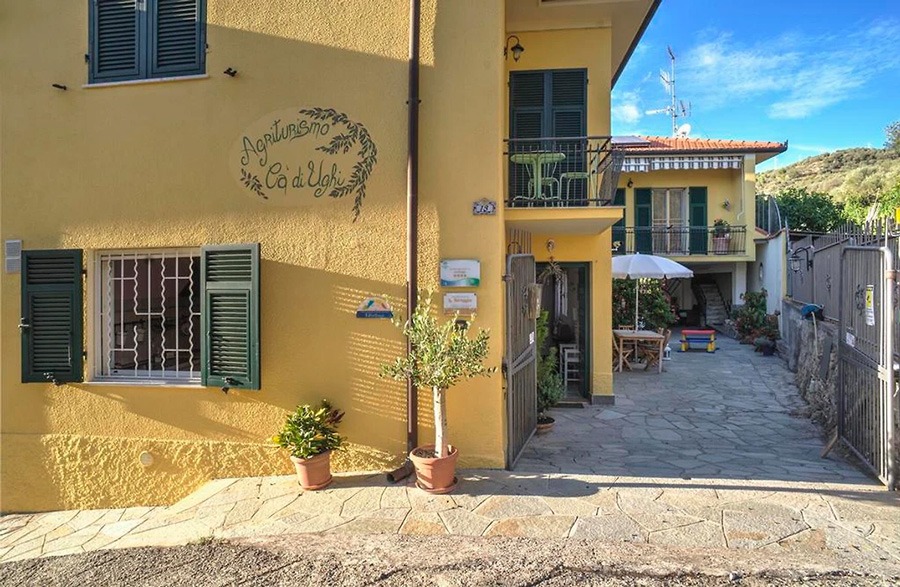 Gelbes Gebäude mit grünen Fensterläden, Balkon und Hofeingang des Agriturismo Cà di Ughi in Diano San Pietro, mit Tischen, Pflanzen und Tor.