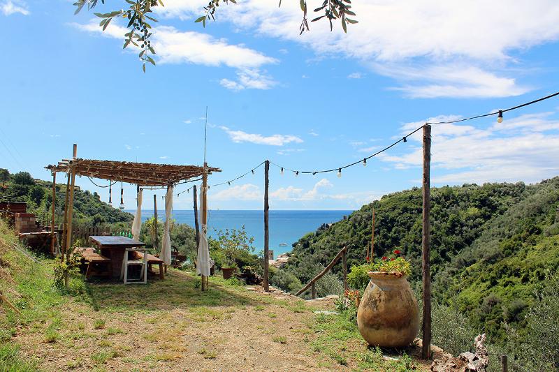 Blick vom Agriturismo Il Brucaolivo auf das blaue Meer von Monterosso al Mare, mit Holzpergola, rustikalem Tisch und grünen Hügeln im Vordergrund.
