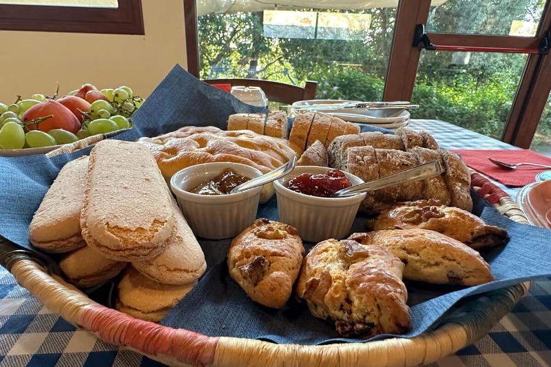 Korb mit Gebäck, Brot, Marmelade und Obst auf einem Tisch am Fenster, dahinter grüner Garten.