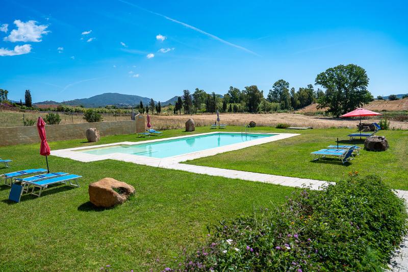 Pool auf einer grünen Wiese mit blauen Liegen und roten Sonnenschirmen, dahinter Felder und niedrige Hügel.
