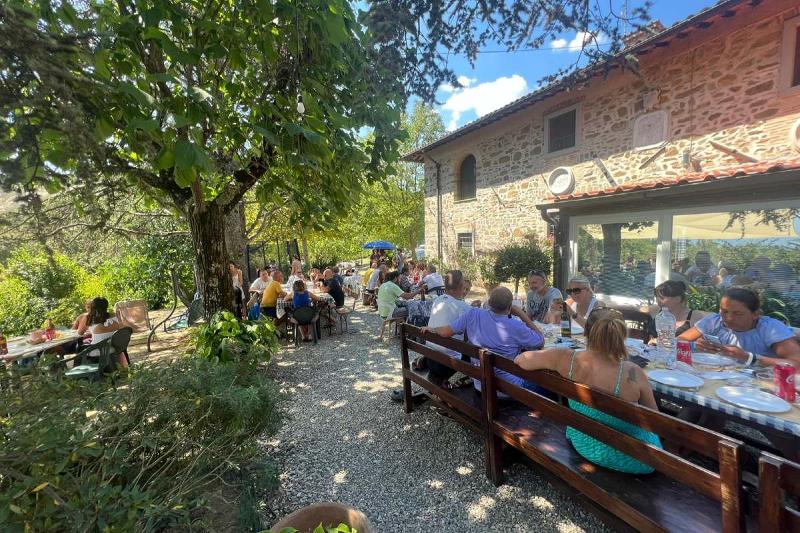 Gäste genießen ein gemeinsames Mittagessen an langen Holztischen im schattigen Garten des Agriturismo Podere Badia ValDrago. Im Hintergrund das restaurierte Steinhaus, umgeben von viel Grün und sommerlicher Atmosphäre.
