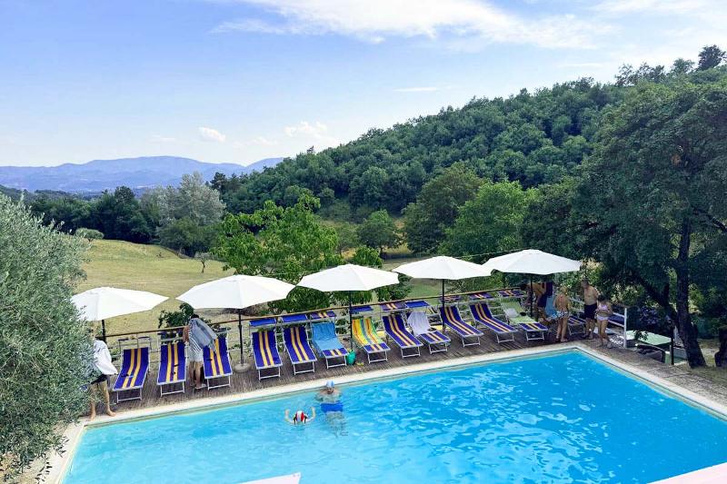 Barrierefreier Swimmingpool auf dem Agriturismo Podere Badia ValDrago in der Toskana, umgeben von Liegestühlen und Sonnenschirmen. Im Hintergrund grüne Hügel und ein weiter Blick in die Landschaft, Menschen entspannen am Pool.